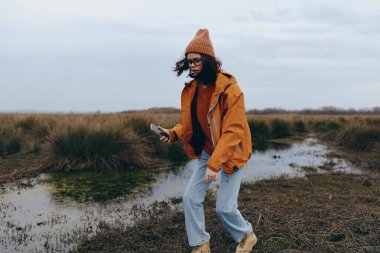 Açık havada, turuncu ceketli ve kahverengi bereli, su kenarında yürüyen ve telefonu tutan bir kadının görüntüsü. Bulutlu hava ortamında günlük sonbahar modası ve doğa macerası.