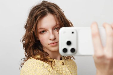 Sarı kazak giyen, bol saçlı bir kızın cep telefonuyla selfie çektiği yakın plan. Modern iletişim, gençlik kültürü ve dijital teknoloji kullanımı kavramı.