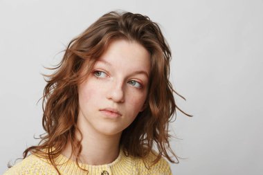 Sarı kazak giyen kıvırcık saçlı ve çilli genç bir kadının portresi, beyaz bir arka planda düşünceli bir ifadeyle başka yere bakıyor. Doğal güzellik ve içgörü kavramı.