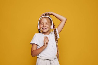Uzun örgülü mutlu genç kız kulaklıklarla müzik dinliyor. Gözleri kapalı, sarı arka planda, günlük beyaz giysiler içinde. Neşe ve çocukluk kavramı.