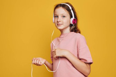 Pembe kulaklık takan ve pastel gömlek giyen gülümseyen genç kız müzikten hoşlanıyor. Kulaklık telini tutuyor ve sarı arka plandaki kameraya bakıyor. Gençlik yaşam tarzı ve teknolojisi kavramı.