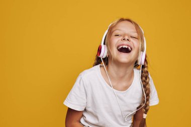 Beyaz tişörtlü gülümseyen genç kız kulaklıkla müzik dinliyor. Parlak sarı zemin mutluluğu, çocukluğu ve neşeli duyguları vurguluyor. Canlı renklerle çekilen stüdyo görüntüleri.
