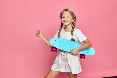 Saçları örgülü mutlu bir kız pembe bir arka planda dikilirken mavi bir kaykay taşıyor. Günlük kıyafet ve kendine güveni olan duruş neşeli, gençlik havası yaratır..