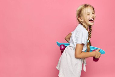 Beyaz elbiseli, örülmüş saçlarıyla gülümseyen çocuk elinde mavi bir kaykayla, kapalı gözlerle gülüyor..