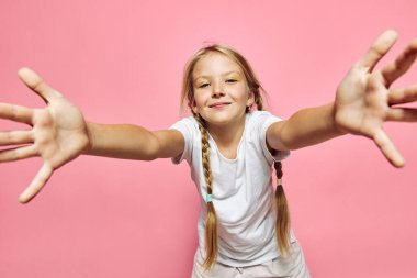 Beyaz gömlekli uzun örgülü gülümseyen genç kız ellerini pembe bir arka fona doğru uzatıyor. Parlak stüdyo ışıkları mutluluğu, eğlenceyi ve masumiyeti vurguluyor..