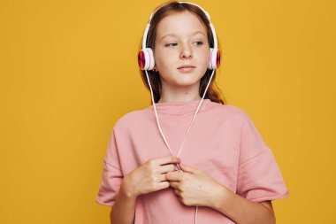 Beyaz kulaklık takan ve pembe gömlek giyen genç kız müzikten hoşlanıyor. O, parlak sarı arka planda sakin bir ifadeyle yana bakıyor. Yaşam tarzı konsepti için stüdyo çekimi.