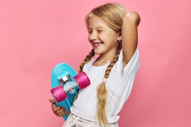 Beyaz tişörtlü gülümseyen sarışın kız pembe tekerlekli renkli bir kaykay tutuyor. Gözleri kapalı ve kaygısız görünüyor. Pembe arka planda stüdyo çekimi.