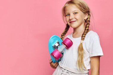 Uzun sarı örgülü mutlu genç kız pembe tekerlekli mavi bir kaykay tutuyor. Beyaz gömlek ve bej pantolon giyiyor, pembe arka planda güvenle duruyor..