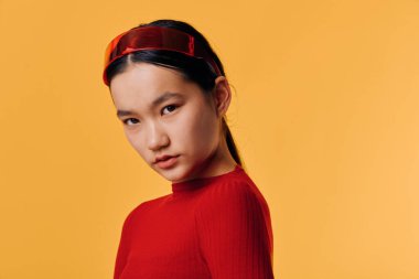 Kırmızı kazak ve saç bandıyla poz veren kısa boylu, uzun siyah saçlı genç bir kadın. Sıcak sarı arka planlı stüdyo portresi ve kameraya özgüvenle bakan doğal stil..