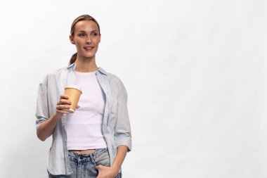 Günlük giysiler içinde mutlu bir kadın tek kullanımlık kahve fincanı tutuyor. Parlak beyaz arka planlı stüdyo çekimi. Sabah rutini, ferahlama ve günlük tarz kavramı.