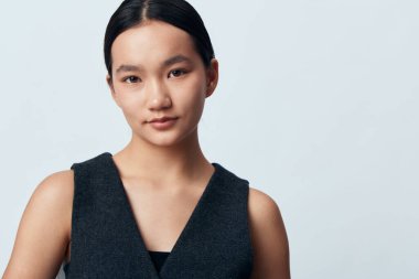 Düz siyah saçlı, doğal makyajlı, kolsuz koyu renk elbisesiyle arka planda kameraya bakan bir kadın model. Güzellik, portre, moda, özgüven, stüdyo çekimi.