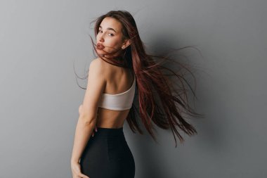 Uzun kahverengi saçlı, beyaz bluzlu ve siyah taytlı genç bir kadının portresi. Moda ya da yaşam tarzı konsepti için tarafsız gri zeminde saçları hareket halinde poz veren kadın.