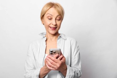 Kısa sarı saçlı, gülümseyen yaşlı kadın akıllı telefon kullanıyor, beyaz arka planda günlük beyaz gömlek giyiyor. Teknoloji, iletişim, mutluluk ve kıdemli yaşam tarzı kavramı.