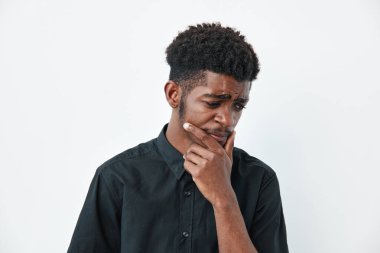 Koyu tenli, siyah gömlekli, elleri çenesinde ve ciddi yüzüyle beyaz arka planda duran Afrikalı bir adam. Derin düşüncelere dalmış genç siyah erkek portresi.