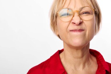 Orta yaşlı, kısa sarı saçlı, gözlüklü ve kırmızı gömlekli bir kadının portresi beyaz arka planda tuhaf bir ekşitme yapıyor. Mizah, duygu ve kişilik kavramı.