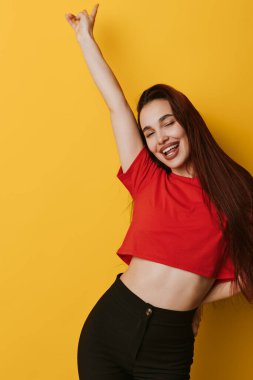 Kırmızı tişörtlü, uzun saçlı, siyah pantolonlu, kollarını kaldırmış poz veren mutlu bir kadın ve sevinç, özgürlük ve pozitif duygu için sarı arka planda kapalı gözler..