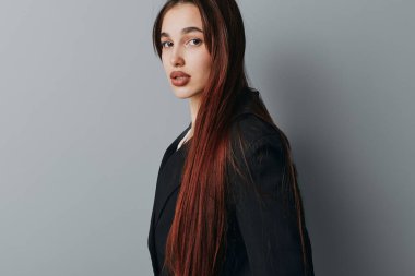 Siyah ceketli, uzun kahverengi saçlı genç bir kadının portresi. Sade gri arka planda sakin bir yüzle dikilip ileriye bakıyor. Sıradan giyinen ve ciddi görünüşlü bir kadının stüdyo fotoğrafı..