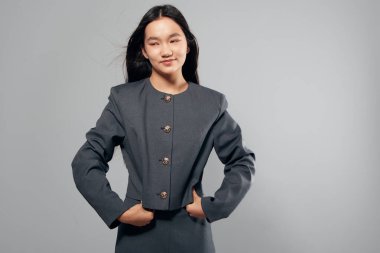 Dekoratif düğmeleri olan gri iş kıyafeti giyen kendine güvenen Asyalı kadın elleri ceplerinde duruyor. Doğal ifadeyle poz veren profesyonel bir kadın model ve açık arkaplanda uzun saçlı..