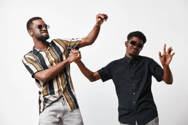 Siyah tenli, günlük kıyafetler ve güneş gözlüğü takmış siyah adamlar beyaz arka planda dans ve arkadaşlığın tadını çıkarıyorlar. Olumlu ruh hali, eğlence ve iki genç erkek arkadaş arasındaki bağ.