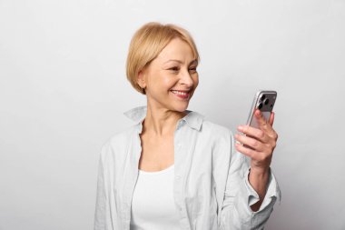 Beyaz günlük giysiler içinde mutlu olgun bir kadın akıllı telefon tutuyor ve gülümsüyor, ekrana bakıyor. Parlak arka plana sahip stüdyo portresi, mobil aygıt konsepti, bağlantı ve neşe.