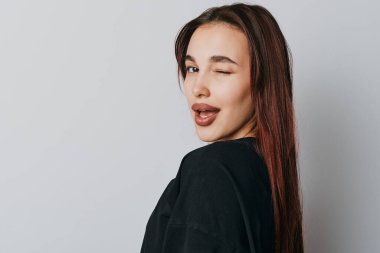 Siyah saçlı çekici genç bir kadın göz kırpıyor ve gri arka planda poz veriyor. Modern fotoğraf çekiminde doğal makyajlı yakın plan portre..