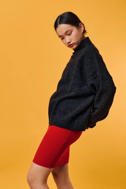 Siyah örgü süveter ve kırmızı etekli genç bir kadın stüdyoda parlak turuncu arka planda poz veriyor. Günlük kıyafetler, minimalist stil, ve moda veya yaşam tarzı çekimi için düşünceli ruh hali..