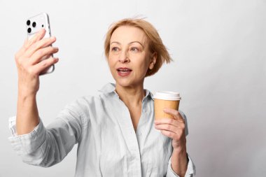 Elinde tek kullanımlık kahve fincanı olan beyaz bluzlu bir kadın beyaz arka planda cep telefonuyla selfie çekiyor. Sosyal medya, iletişim, yaşam tarzı ve teknoloji kavramı.