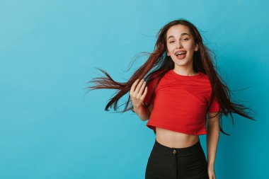 Kırmızı tişörtlü, koyu renk pantolonlu, mavi duvarın önünde poz veren mutlu kız doğal görünüş ve tasasız tavırla neşeyle gülümsüyor. Gençlik enerjisi ve renkli moda konsepti.