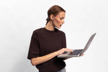 Gündelik koyu renk kazaklı genç bir kadın iki eliyle dizüstü bilgisayar tutuyor ve beyaz arka planda geniş gözlerle ekrana bakıyor. Dijital iletişim ve çevrimiçi tepki kavramı.