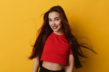Kırmızı gömlekli, uzun kahverengi saçlı, parlak sarı duvarda poz veren mutlu kız. Doğal gülüşü olan ve yaşam tarzı, güzelliği ve gençlik konsepti için günlük kıyafetleri olan bir kadın model..