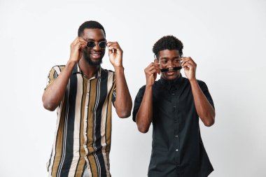 Siyah tenli iki Afrikalı Amerikalı adamın portresi güneş gözlüğü takıyor ve moda ve arkadaşlık teması için beyaz arka plana karşı günlük kıyafetler içinde gülümsüyor..