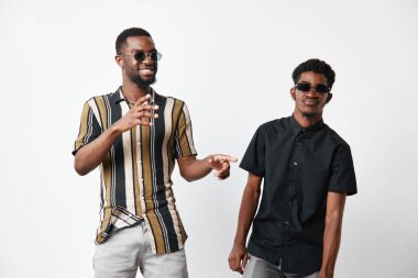 Siyah tenli, günlük giysiler ve güneş gözlüklü Afro-Amerikalı erkekler mutlu ifadeli ve rahat vücut dili olan bir yaşam tarzı portresi için beyaz arka planda poz veriyorlar..