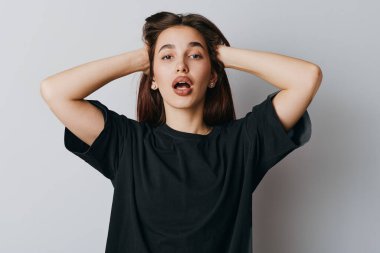 Kahverengi saçlı, siyah tişörtlü, ağzı açık, el ele tutuşmuş, beyaz arka planda yaşam tarzı ve ruh hali için tarafsız bir yüz ifadesi olan sıradan bir genç kadın..