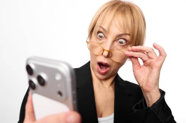 Akıllı telefonu kapalı tutan, ağzı açık, ağzı açık, beyaz arka planda siyah ceket giyen olgun bir kadın. Sürpriz, tepki, dijital iletişim kavramı.