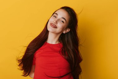 Kırmızı tişörtlü, uzun siyah saçlı gülümseyen kız sarı arka planda rahatça poz veriyor, doğal makyajlı rahat bir portre ve parlak stüdyo ortamında rahat bir ruh hali..