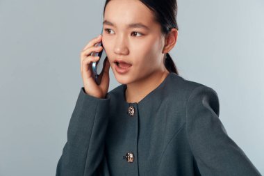 Açık ağızlı Asyalı bir kadın telefonuna tepki veriyor. Tarafsız bir arkaplan önünde spor ceket giyiyor. Kadın cep telefonunu iletişim, müşteri hizmetleri ya da modern kıyafette sosyal sohbet için kullanıyor ve...