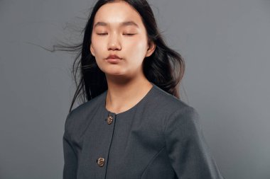 Uzun siyah saçlı, gözleri kapalı, resmi gri elbiseli genç bir manken. Yumuşak ışıklandırma, nötr gri arkaplanda portre fotoğrafı için pürüzsüz cilt ve gevşek yüzü vurgular.