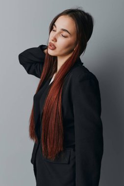 Uzun örgülü, sakin yüzlü, siyah ceketli ve beyaz bluzlu, elleri gri arka planda duran genç bir kadının portresi..