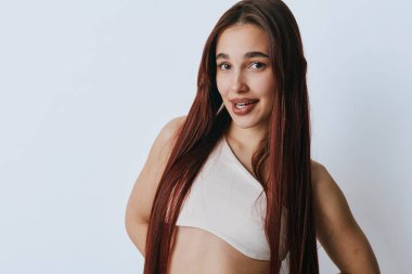 Uzun düz kızıl saçlı, beyaz kolsuz bluzlu, beyaz arka planda poz veren kendine güvenen genç bir kadın. Doğal görünüşlü ve sıradan bir tarzı olan kadın model kameraya hafifçe gülümsüyor..