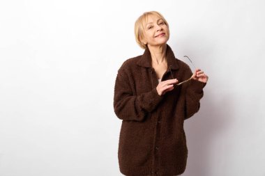 Kahverengi ceketli, gülümseyen yaşlı kadın beyaz arka planda poz veren gözlük tutuyor. Tarz, güven ve olgun güzellik anlayışı. Günlük kıyafet ve rahat ruh hali..