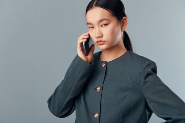 Koyu renk iş ceketli bir kadın iletişim için akıllı telefon kullanıyor. Gri arka plana karşı duruyor. Ciddi yüz ifadesi konsantrasyon ve nişan gösteriyor..