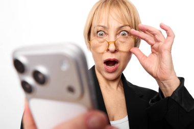 Şok olmuş orta yaşlı gözlüklü kadın akıllı telefonu yanında tutuyor, şaşırmış bir ifadeyle gözlüklerini ayarlıyor. Dijital iletişim, beklenmedik bilgi ve duygusal tepki kavramı.