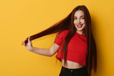 Kırmızı gömlekli, uzun kahverengi saçlı ve sarı arka planda duran siyah pantolonlu gülümseyen kız. Doğal görünüşlü neşeli bir kadın portresi ve saçı olan neşeli bir jest..