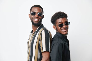 Siyah tenli, güneş gözlüğü ve günlük kıyafetlerle sırt sırta poz veren Afro-Amerikalı erkekler. Gülümseyen genç erkekler, modaya uygun saç stilleri ve rahat bir duruşu var..