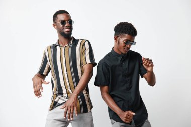 Siyah tenli, güneş gözlüğü takan ve beyaz arka planda mutlu dans eden iki siyah adam. Genç arkadaşlar neşeli ifadelerle eğleniyor ve eğleniyorlar..