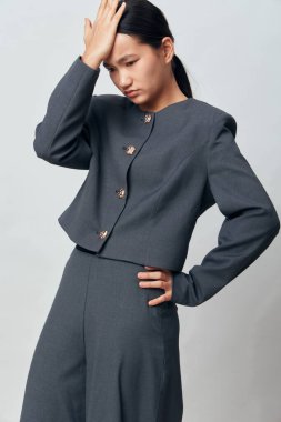 Düğmeleri olan gri takım elbiseli genç bir kadının portresi, düşünceli duruşu, uzun koyu renk saçı, profesyonel kıyafeti, temiz bir geçmişi ve kendine güveni olan bir duruşu var..