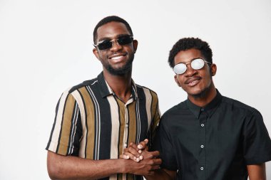 Güneş gözlüklü iki esmer adam el sıkışıyor ve gülümsüyor. Beyaz stüdyo arka planında pozitif enerjili gündelik tarz, arkadaşlık ve mutluluk.
