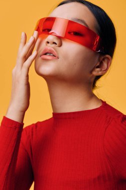 Parlak kırmızı bluzlu ve şeffaf kırmızı gözlüklü genç bir kadın yüzüne kapalı dudaklarla dokunuyor. Portre fotoğrafçılığı cesur renkli görünüş ve turuncu arka planda yaratıcı moda aksesuarı..