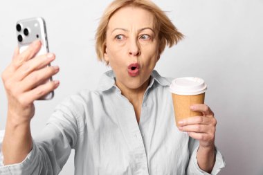 Kısa saçlı, tek kullanımlık kahve fincanı tutan olgun bir kadın beyaz arka planda telefonla selfie çekiyor. Teknoloji, ifade, sosyal medya ve günlük rutin kavramı.