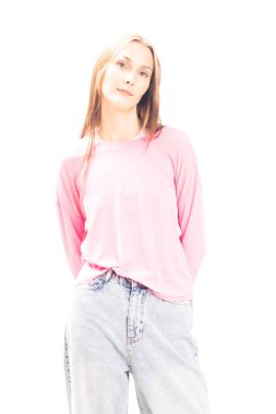 Beyaz arka planda poz veren pembe kazaklı ve kot pantolonlu genç bir kadının portresi. Parlak stüdyo ışığı doğal ten rengini ve basit modern tarzı ön plana çıkarıyor.